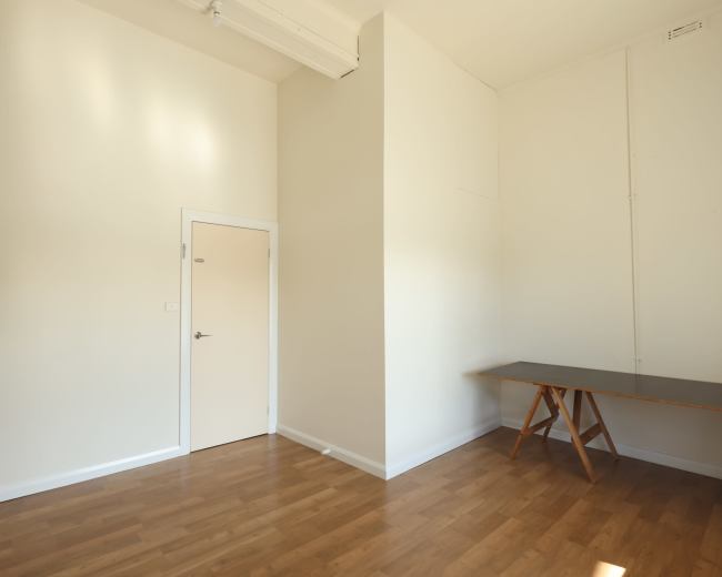 The image shows a minimalist room with light-colored walls, a wooden floor, a plain door, and a wooden table against one wall.