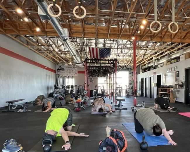 A group of people performs exercises on mats in a spacious gym with weights and fitness equipment visible in the background.