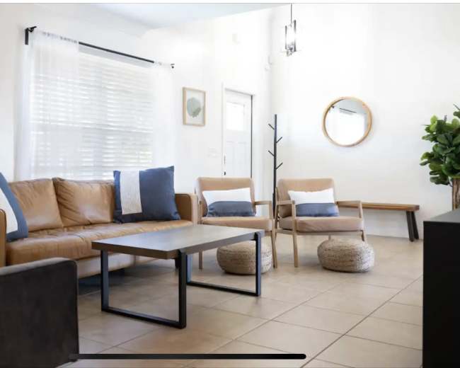 A modern living room with a tan leather sofa, two wooden chairs, a coffee table, and a round mirror on the wall.