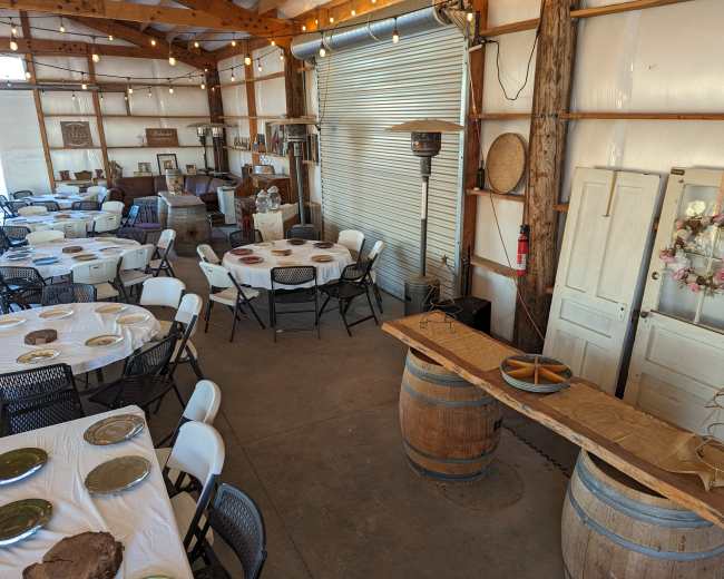 A spacious indoor setting with tables covered in white tablecloths arranged around a central area, featuring decorative elements like string lights, barrels, and old wooden doors.