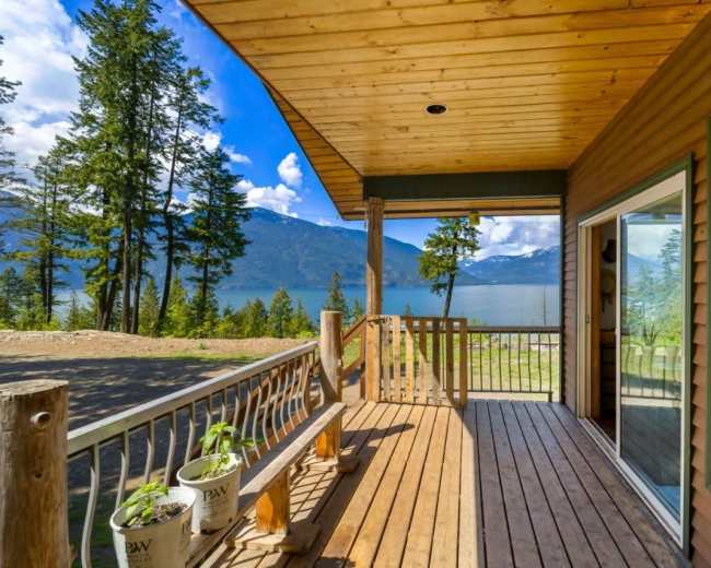 The image shows a wooden deck overlooking a lake, surrounded by tall trees and distant mountains under a partly cloudy sky.