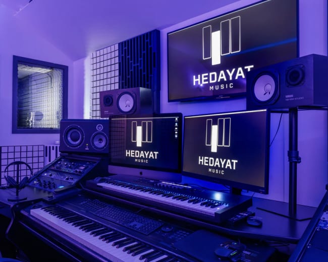 The image shows a modern music studio featuring a keyboard, multiple monitors displaying "HEDAYAT MUSIC," and professional audio equipment in a dimly lit room with blue lighting.