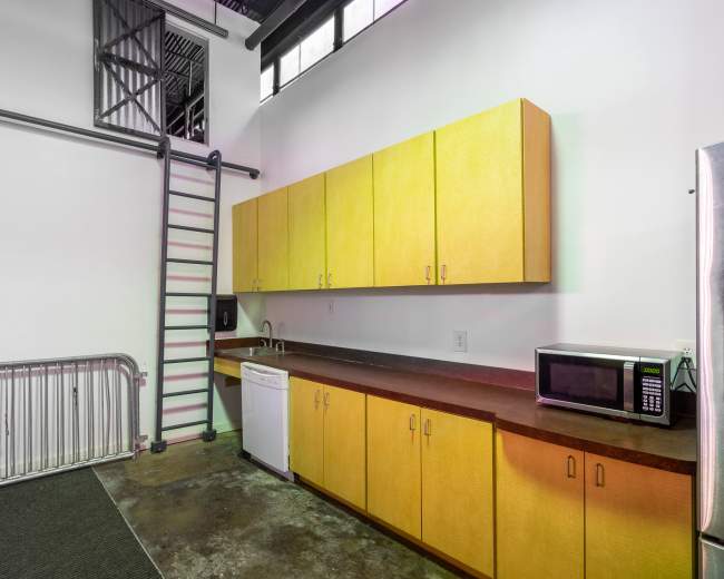 The image shows a minimalist kitchen with yellow cabinetry, a microwave, a dishwasher, and a metal ladder leading to a loft area.