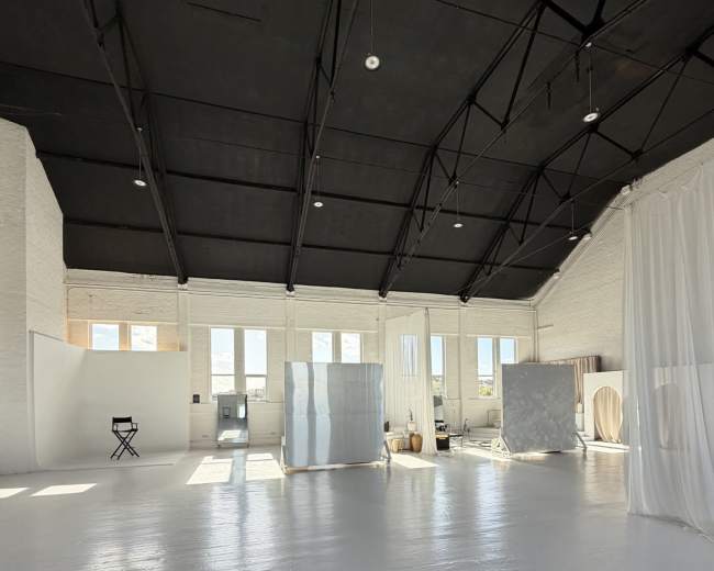 The image shows a spacious, well-lit studio with large windows, white walls, and several fabric dividers creating distinct areas.