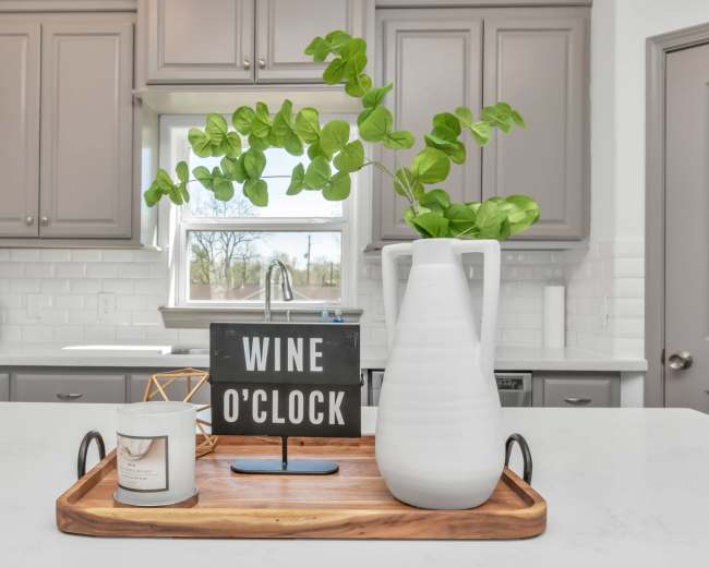 A wooden tray on a kitchen countertop displays a candle, a decorative object, and a vase with green foliage, alongside a sign that reads "WINE O'CLOCK."