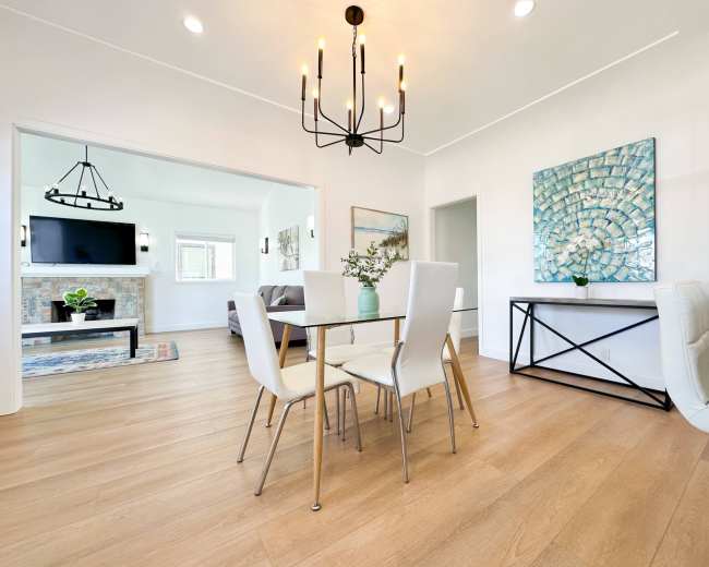 The image shows a modern dining area with a glass-top table and white chairs, featuring a chandelier and a decorative wall art piece.