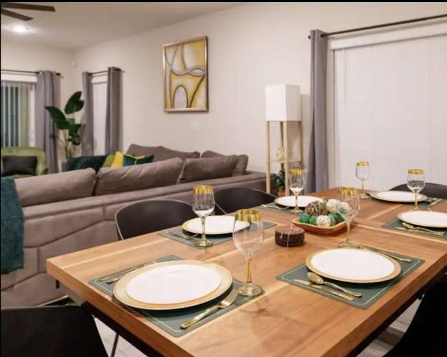 A dining table is set for six with plates, glasses, and decorative items in a modern living space featuring a comfortable gray sofa and plant accents.