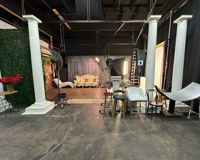 The image shows a photography studio with decorative columns, various chairs, and backdrop setups.