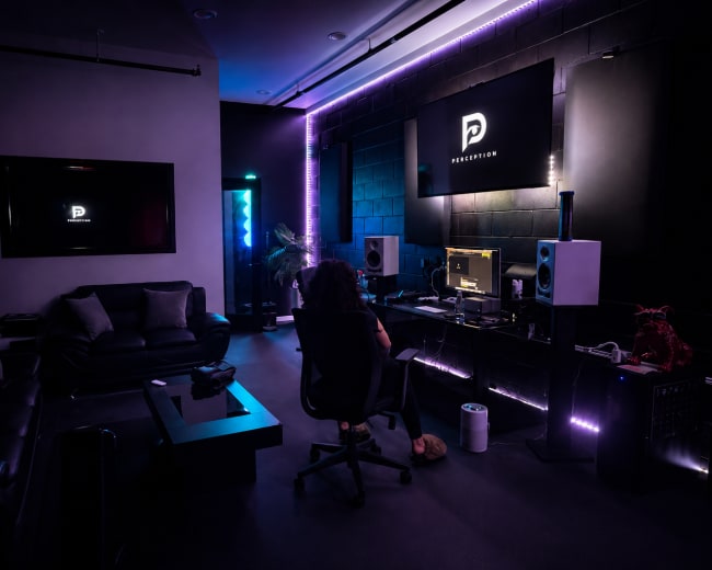 A person sits at a desk in a dark room filled with sound equipment and ambient lighting, working on a music production setup.