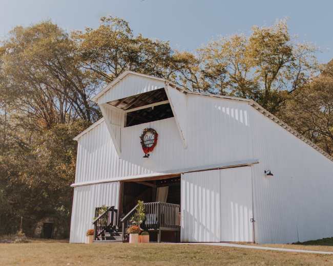 A white barn with a large opening at the top and decorative elements stands on a grassy area surrounded by trees.