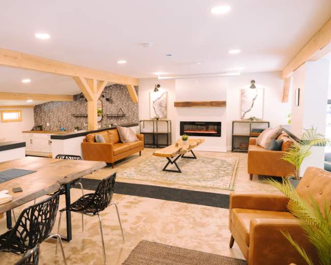 The image depicts a modern, open-concept living area featuring a kitchen, dining space, and seating area with wooden beams and a central coffee table.