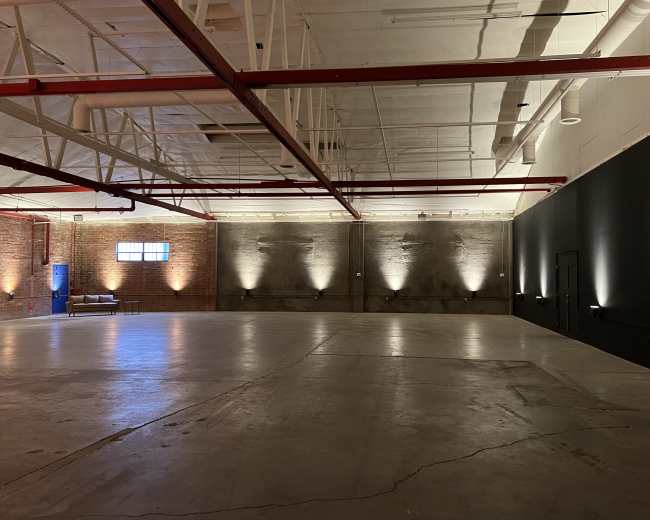 The image shows a spacious, empty industrial-style room with exposed brick walls, a concrete floor, and recessed lighting along one wall.