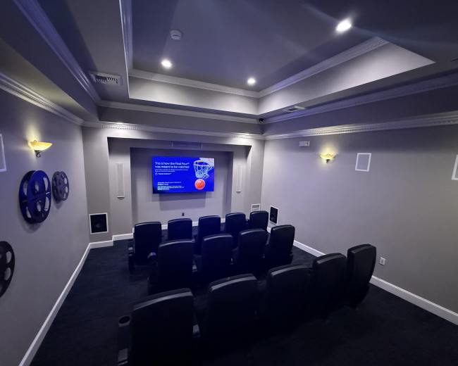 The image shows a home theater with tiered seating, a large screen displaying a presentation, and decorative film reel wall art.