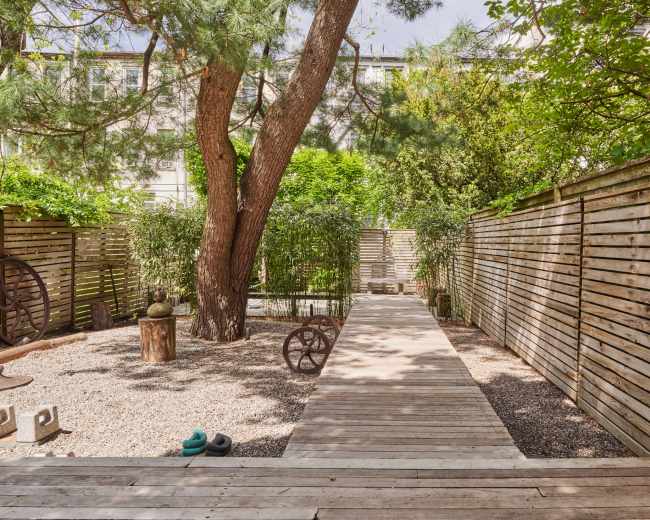 A wooden pathway runs between tall trees and wooden fences in a landscaped garden.