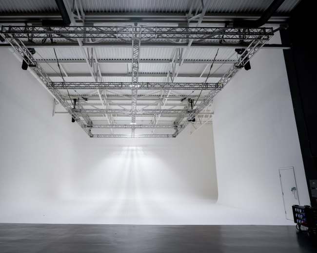 The image shows a spacious, well-lit photography studio with a white backdrop and overhead lighting apparatus.