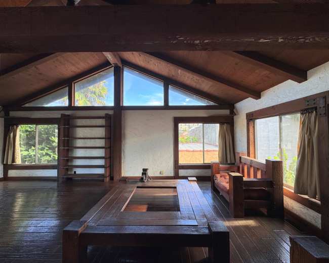 The interior of a wooden cabin features high ceilings, large windows with natural light, and minimalist furnishings including a wooden table and bench.