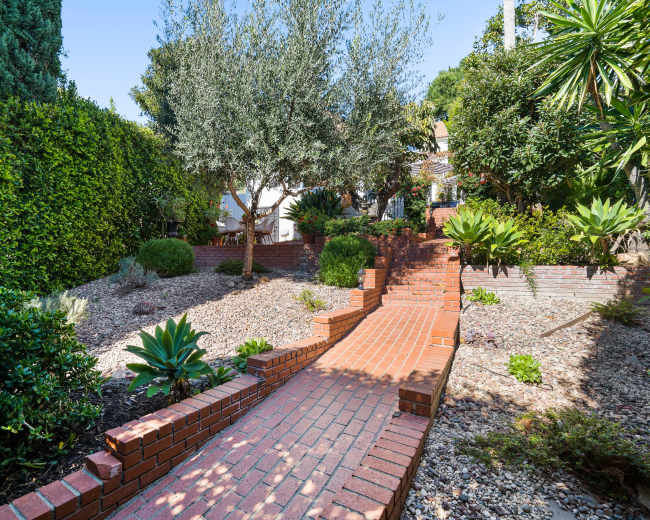 A brick pathway leads through a landscaped garden featuring shrubs, small trees, and decorative stones.