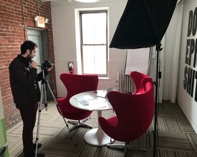 A person is setting up a camera on a tripod in a room with red chairs and a motivational message on the wall.