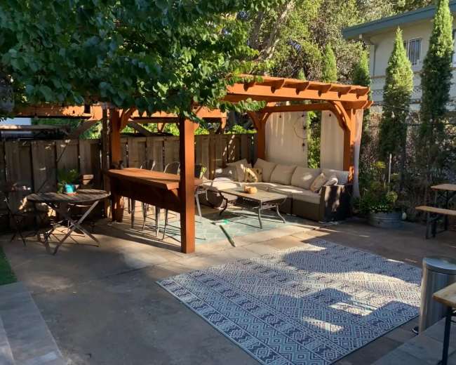 The image shows a backyard patio with a wooden pergola, furniture arranged for seating, and a rug on the concrete floor.