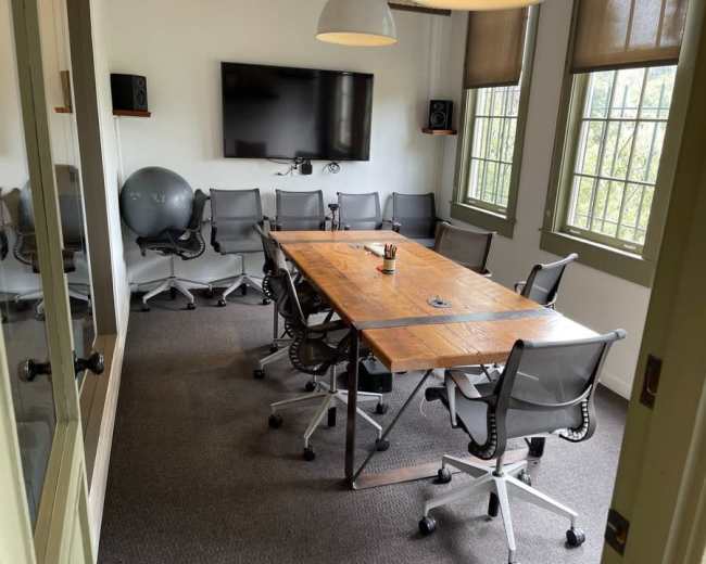 The image shows a conference room with a large wooden table surrounded by several ergonomic chairs, a large screen mounted on the wall, and a large exercise ball in one corner.
