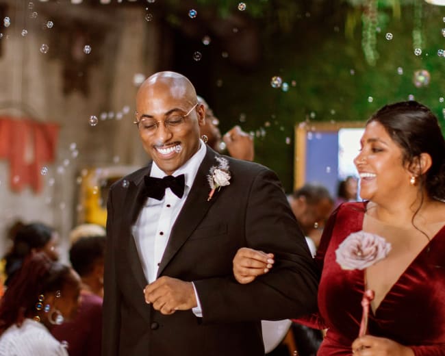 A man in a tuxedo and a woman in a red dress walk together among guests celebrating with bubbles at a wedding.