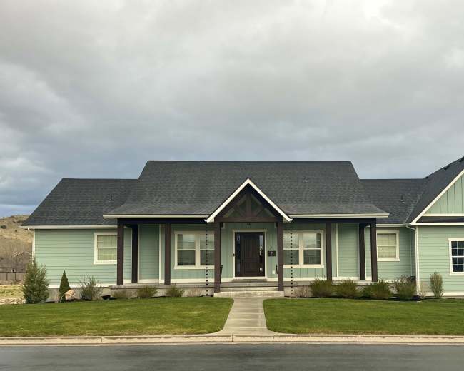 The image shows a large, light green house with a dark roof and a well-maintained lawn, situated on a residential street.