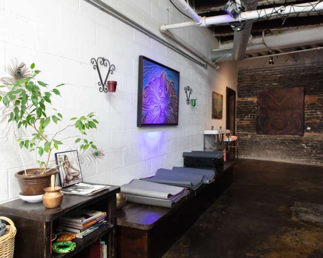 The image shows an art studio with a wooden bench displaying several yoga mats, framed artwork on the walls, and exposed brick.