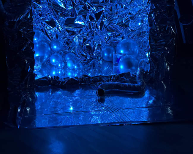 The image features a reflective, metallic backdrop illuminated in blue, with spherical objects and tubes arranged in front of it.