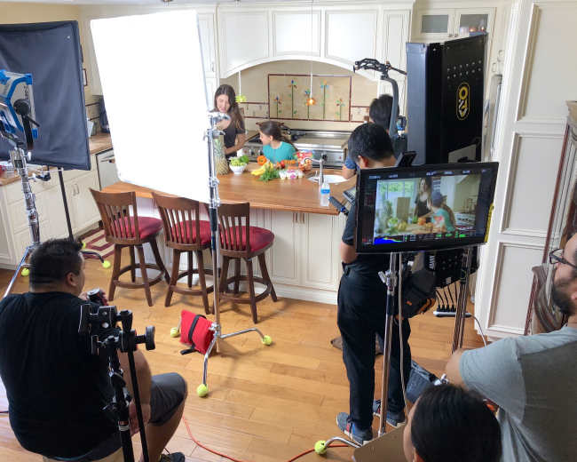 A film crew is set up in a kitchen, with lights and cameras focused on a woman and child at the counter preparing food.