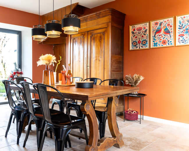 The dining area features a wooden table surrounded by black chairs, with a brown-painted wall and large windows allowing natural light to enter.