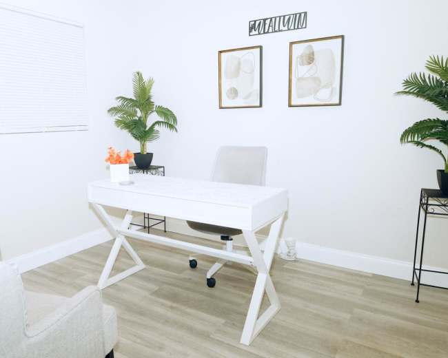 The image shows a minimalist office space featuring a white desk with a chair, two potted plants, and framed wall art.
