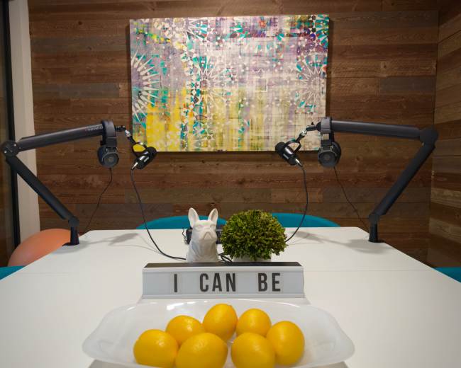 A brightly lit room features a table with a bowl of lemons, a decorative plant, and a sign reading "I CAN BE," against a colorful abstract painting and two microphone stands.