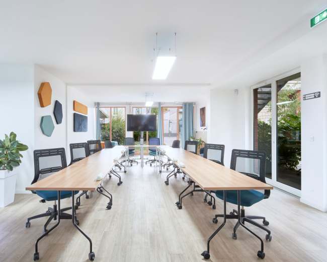 A modern conference room features a long table surrounded by ergonomic chairs, with large windows providing natural light and greenery visible outside.