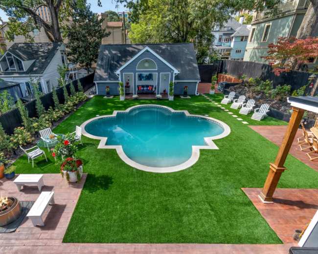 The image shows a backyard featuring a green synthetic lawn with a pool in the shape of a quatrefoil and seating areas surrounded by decorative plants.