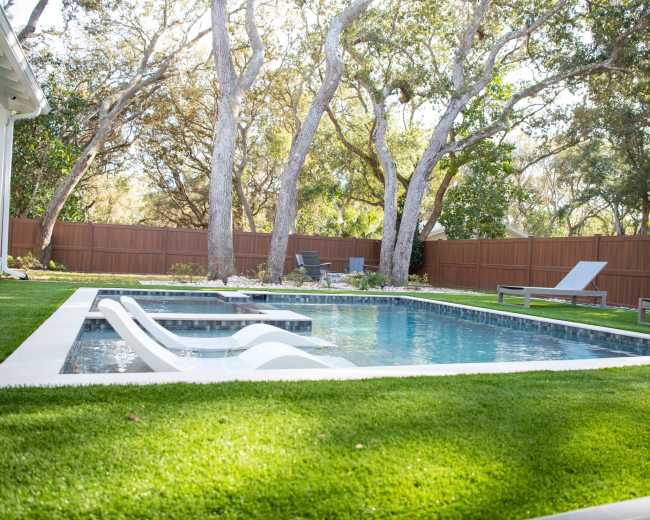 The image shows a rectangular swimming pool surrounded by artificial grass and trees in a backyard setting.