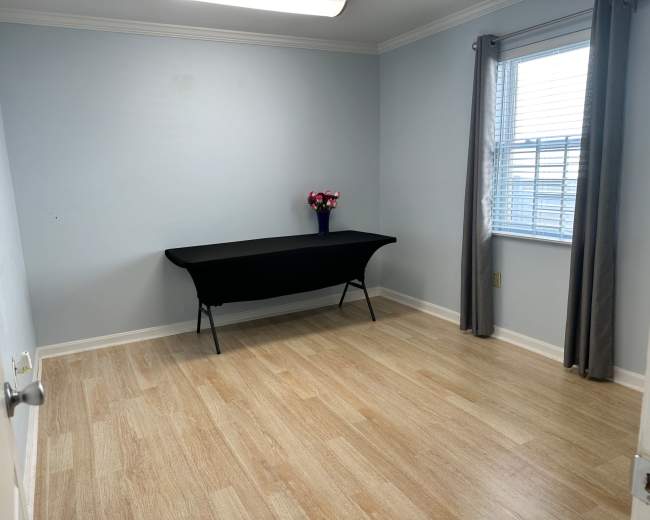The image shows a light blue room with a wooden floor, featuring a black table in the center and a window with sheer curtains.