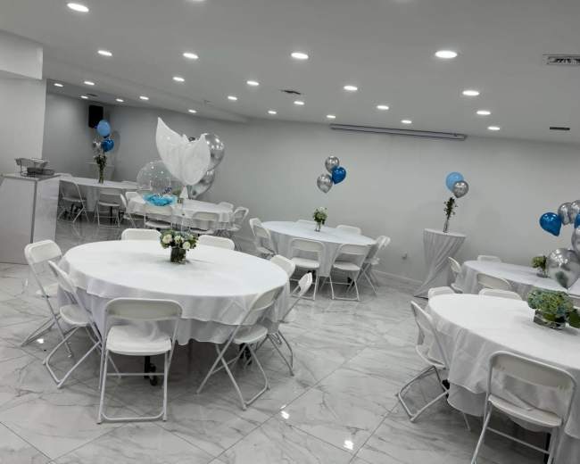 A brightly lit event space with several round tables covered in white tablecloths, decorated with centerpieces and surrounded by balloons.
