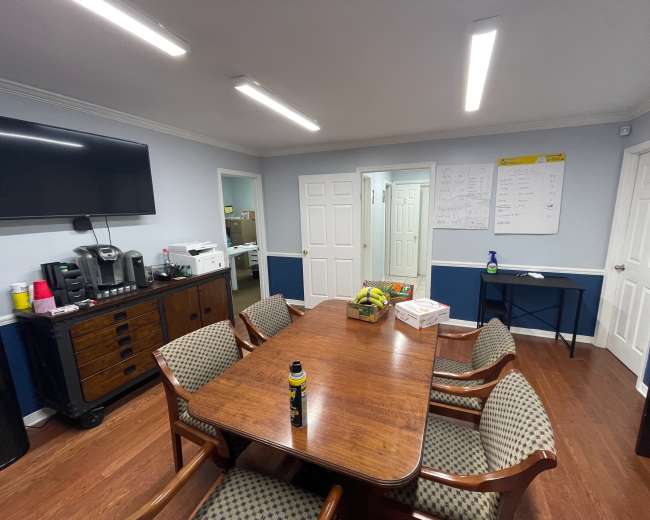 A conference room features a wooden table surrounded by chairs, a television mounted on the wall, and a coffee station with supplies nearby.