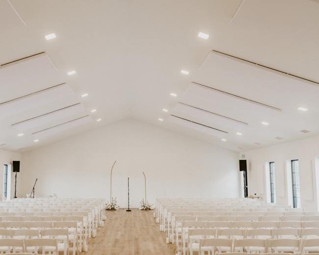 The image shows a spacious, well-lit indoor venue set up with rows of white folding chairs facing a central stage area with decorative elements.
