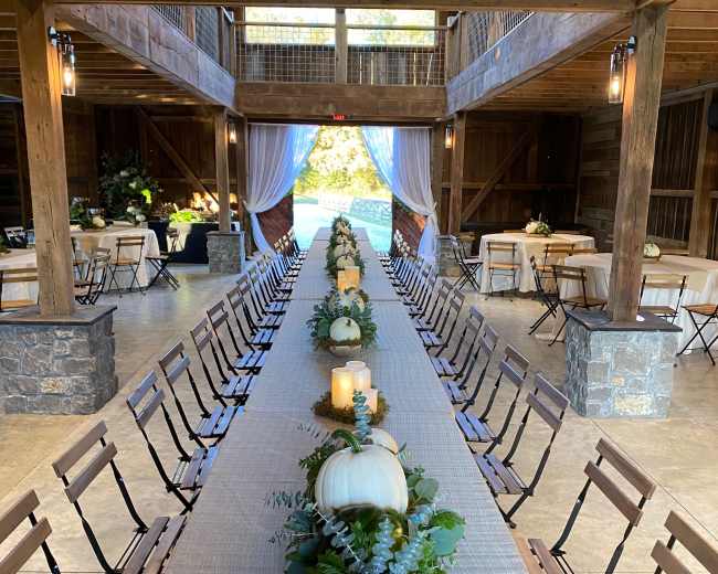 A long dining table is set with white pumpkins and greenery as a centerpiece, placed in a rustic indoor venue with wooden beams and large windows.
