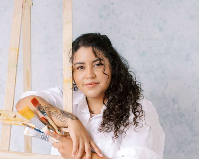 A person with curly hair sits on the floor next to a wooden easel while holding paintbrushes.