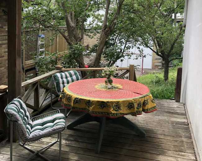A wooden deck features a round table with a vibrant tablecloth and a potted plant at its center, surrounded by trees and greenery.