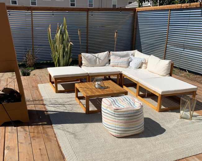 A wooden patio features a sectional sofa with white cushions, a coffee table, a pouf, and a large area rug on a wooden deck.