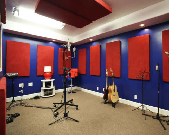 The image shows a sound studio featuring blue walls, red acoustic panels, various microphones on stands, a guitar, and a lamp on a white table.