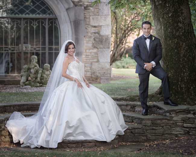 A bride in a white wedding dress and a groom in a black tuxedo pose together in a garden setting near a stone wall and greenery.