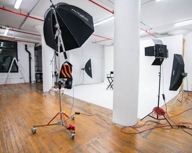 A photography studio is set up with various lighting equipment, a white backdrop, and a director's chair in the center.