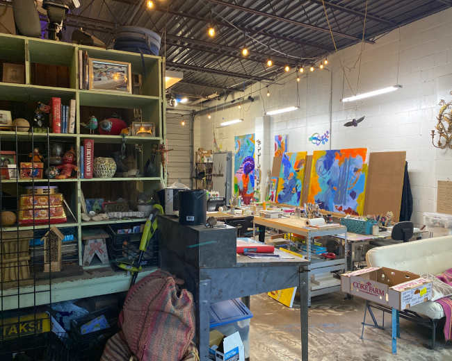 The image shows an art studio filled with various supplies, furniture, and vibrant paintings on the walls.