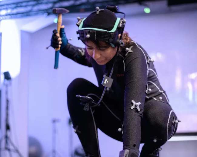 A person in a motion capture suit crouches while holding a hammer above a wooden stick on the ground.