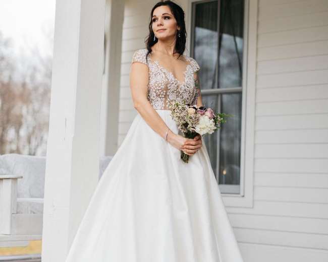 A woman in a wedding dress with a beaded bodice stands outside a white house holding a bouquet of flowers.