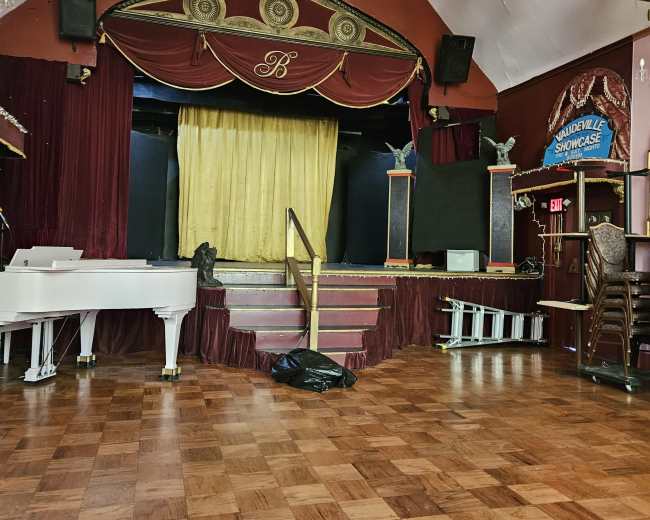 An empty stage with a red and gold curtain, flanked by two decorative pedestals, in a room with wooden flooring and a grand piano on the side.